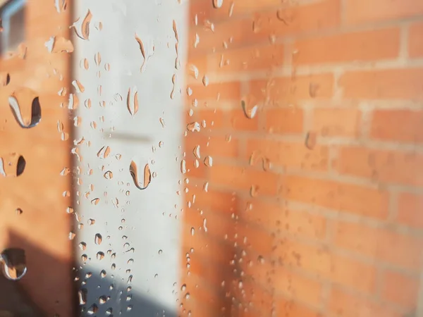 Fenêtre en gouttes de pluie gros plan, une goutte de pluie sur le verre. Mur en brique orange avec porte extérieure comme fond . — Photo