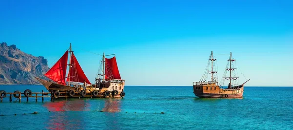 Two old wooden ships. One of them with red sails is on the pier.