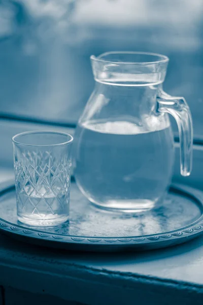 Jarra de agua y un vaso en el alféizar de la ventana, tono azul . — Foto de Stock