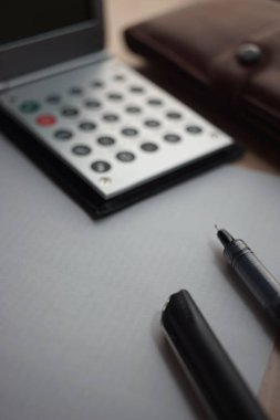 Beyaz boş kağıt üzerinde siyah kalem, metal bir hesap makinesi ve masanın üzerinde bulanık bir deri çanta..