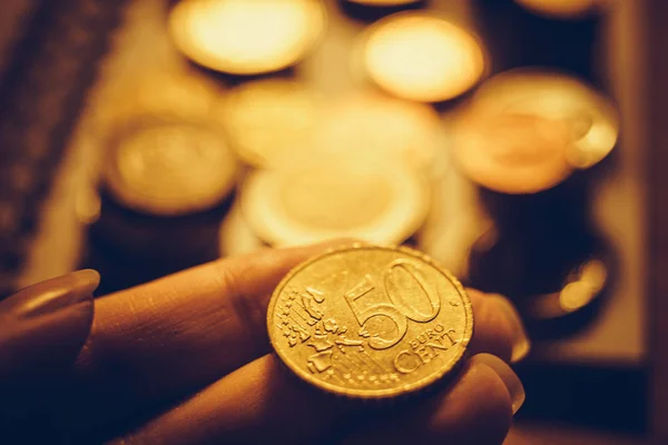 Moneta d'oro da 50 centesimi di euro in mano umana, denaro nelle dita femminili, pile di monete metalliche e tavolo sullo sfondo . — Foto Stock