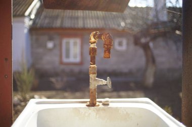 Kırsal bir bahçedeki lavaboda eski paslı bir musluk. Evin ön cephesi arka planda, bulanık. Güneşli bir gün