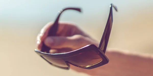 Male hand holds large brown sunglasses. Blurred background of a sunny beach. — Stock Photo, Image