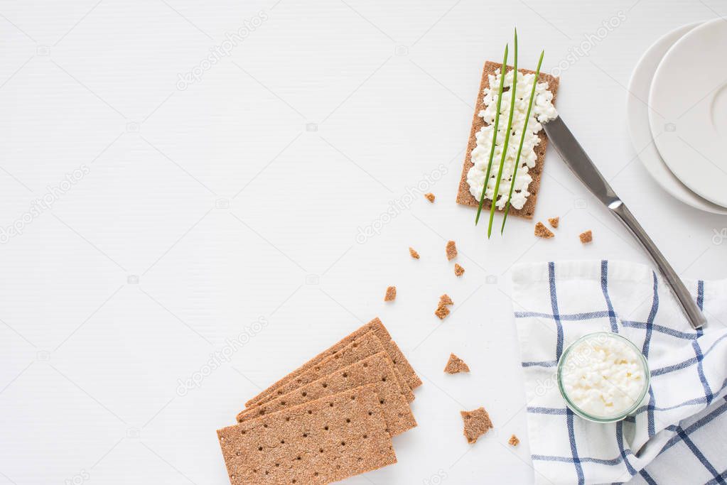 Brown rye crispy bread (Swedish crackers) with spread cottage cheese, decorated with thin green onion, on piece of cloth with plates