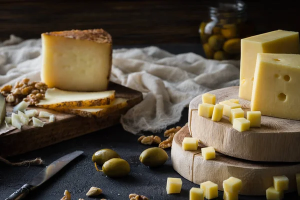 Cheese platter of chopped Swedish hard cheese and sliced Spanish manchego and Italian pecorino toscano on wooden boards, with green olives in glass jar and walnuts — Stock Photo, Image