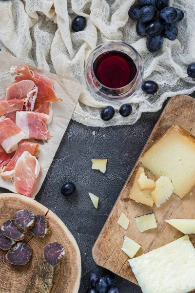Platter with hard cheeses (Italian pecorino toscano and Spanish manchego), homemade dried meat, ham jamon serrano or prosciutto crudo, glass of red wine with grapes, top view on dark rustic background — Stock Photo, Image