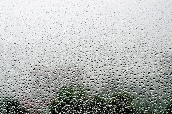 Gouttes de pluie sur verre gros plan. Contexte des arbres et des bâtiments à l'extérieur — Photo