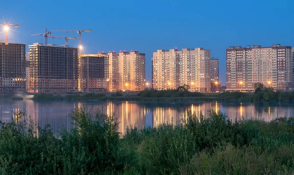 Nytt bostadsområde under uppförande på stranden av floden mot bakgrund av nattljus reflekteras i vattnet med kranar — Stockfoto