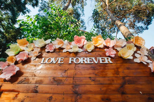 Häät merkintä "rakkaus ikuisesti" puinen seinä kukkia taustalla sininen taivas ja puut — kuvapankkivalokuva