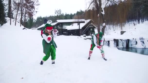 緑の衣装と長いフードのエルフの雪の中で再生されます 冬の背景と2つの幸せな妖精の文字は 森の中で家の前に雪玉を演奏 — ストック動画