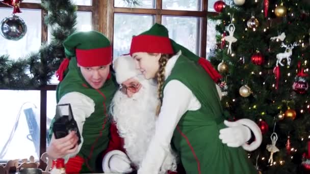 サンタとエルフはクリスマスの装飾の背景で自撮りをしています レトロカメラで写真を撮る妖精 — ストック動画