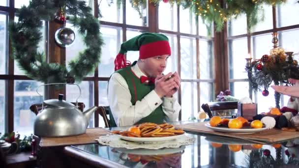 クリスマスの背景にテーブルに座ってカップを持つエルフの男性 面白いです自己で緑の衣装から立ち上がるテーブル — ストック動画
