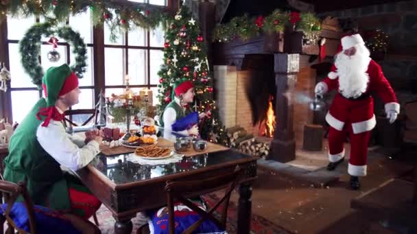 圣诞老人给精灵们倒热水 圣诞装饰背景在室内 童话人物在节日桌上 — 图库视频影像