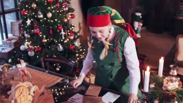 身穿绿色西服的快乐小精灵女孩把信转移到一个房间的桌子上 桌上有圣诞树和装饰品 在壁炉的后面 — 图库视频影像