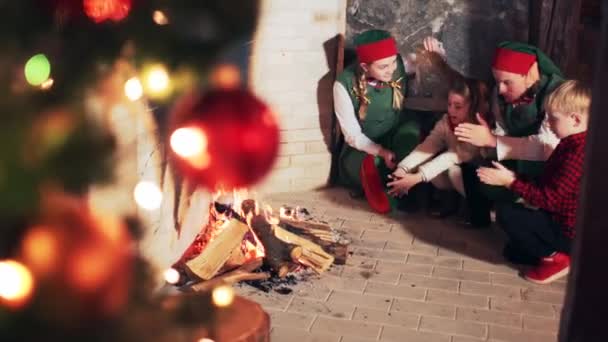 快乐的孩子们和圣诞老人小精灵们用他们的手温暖着房间里的壁炉旁的圣诞树 — 图库视频影像
