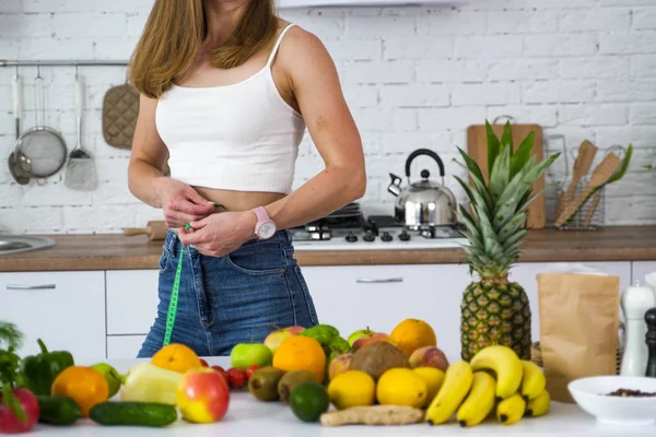 A vegan girl in the kitchen near the fruit measures her waist.