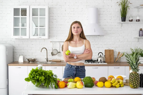 Kaunis tyttö pöydän lähellä, jossa on paljon vihanneksia ja hedelmiä valkoisessa keittiössä, pitää kädessään omenaa. . — kuvapankkivalokuva