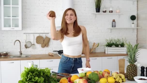 陽気な女の子がココナッツを手に遊んでいる 野菜や果物をテーブルの上に白いキッチン — ストック動画