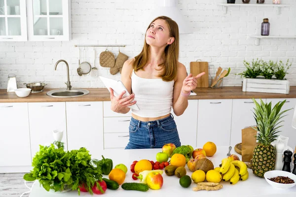 Nuori tyttö keittiössä tabletti käsissään etsii reseptejä ruoanlaittoon. Pöytä, jossa on erilaisia vihanneksia ja hedelmiä terveelliseen kasvisruokavalioon . — kuvapankkivalokuva