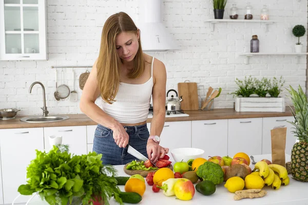 Tyttö leikkaa veitsellä ja tomaatilla. Valkoinen keittiö ja pöytä hedelmiä ja vihanneksia ruoanlaittoon kasvisruokaa . — kuvapankkivalokuva