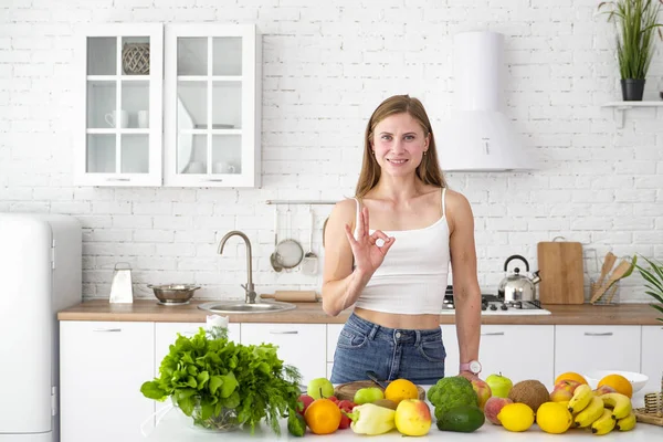 เด็กสาวแสดงสัญลักษณ์โอเคในห้องครัวสีขาว โต๊ะที่มีสมุนไพรผักและผลไม้ โภชนาการเพื่อสุขภาพที่เหมาะสม . — ภาพถ่ายสต็อก