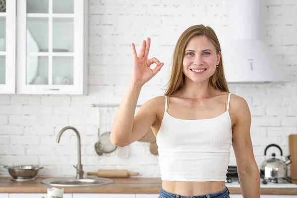 Hymyilevä tyttö näyttää sormin ok. Valkoinen keittiö taustalla. Terveellinen syöminen . — kuvapankkivalokuva