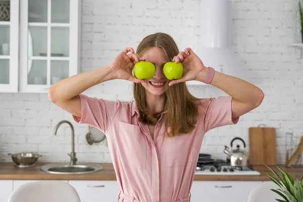 Hymyilevä nuori nainen valkoisessa keittiössä sulkee silmänsä kahdella vihreällä omenalla . — kuvapankkivalokuva