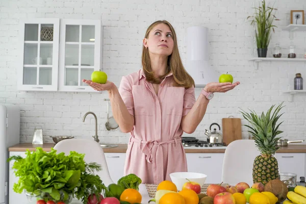 เด็กสาวจินตนาการในครัวสีขาวที่มีผักและผลไม้ถือแอปเปิ้ลสองตัวในมือของเธอ . — ภาพถ่ายสต็อก