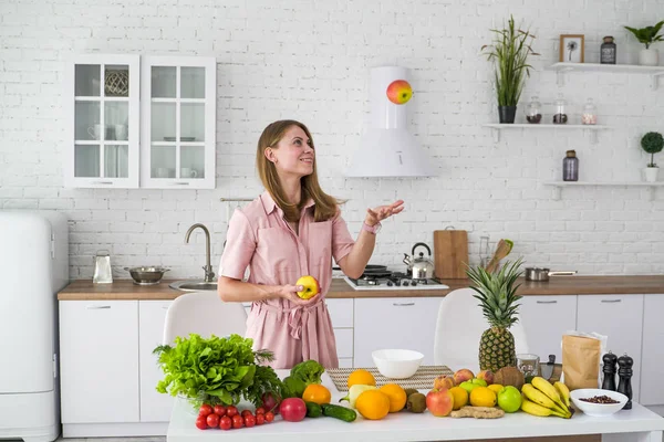 Iloinen tyttö keittiössä lähellä pöytää hedelmiä ja vihanneksia heittää omenan. Terveellinen kasvisruoka . — kuvapankkivalokuva