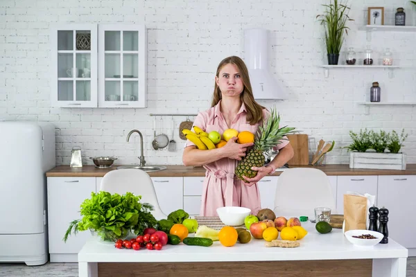 Hauska tyttö keittiössä pitää käsissään sylillinen eksoottisia hedelmiä ja suuri ananas. Valkoinen keittiö ja pöytä erilaisia vihanneksia ja hedelmiä . — kuvapankkivalokuva