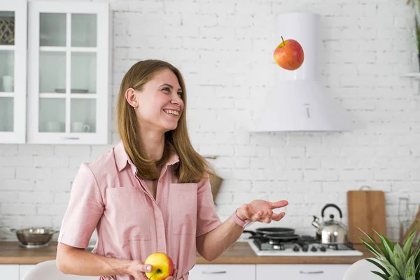 Hymyilevä tyttö heittää punaisen omenan ilmaan valkoisessa keittiössä. Kasvisruoka . — kuvapankkivalokuva