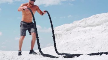 Şortlu atletik bir adam, beyaz bir dağın arka planına karşı doğa üzerindeki iplerle egzersiz yapıyor. Yavaş çekim.