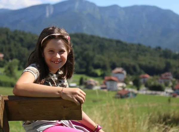 Kaukaski całkiem mała dziewczynka z długimi włosami uśmiechający się na ławce w — Zdjęcie stockowe