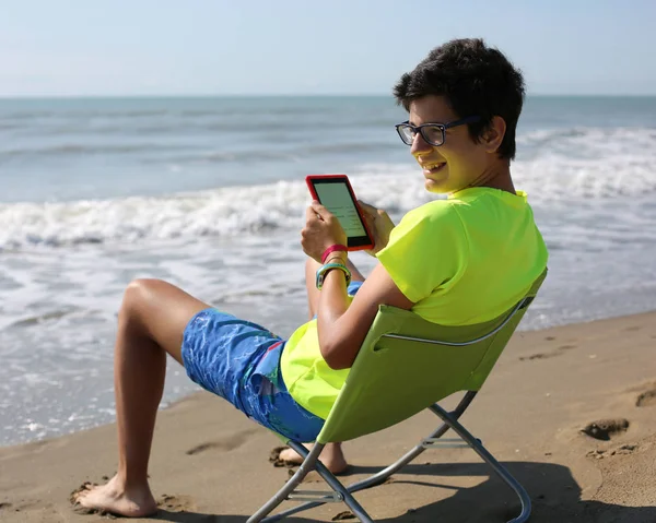ビーチの椅子に座って電子ブックを読む白人少年の笑顔 — ストック写真