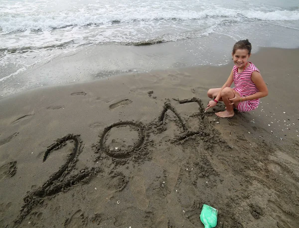 Uśmiechający się mała dziewczynka, w pobliżu morza rysuje piśmie rok 2 — Zdjęcie stockowe