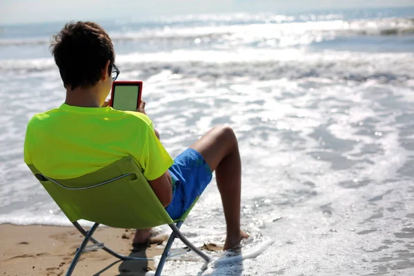 Młody chłopak czyta ebook przez ocean latem — Zdjęcie stockowe