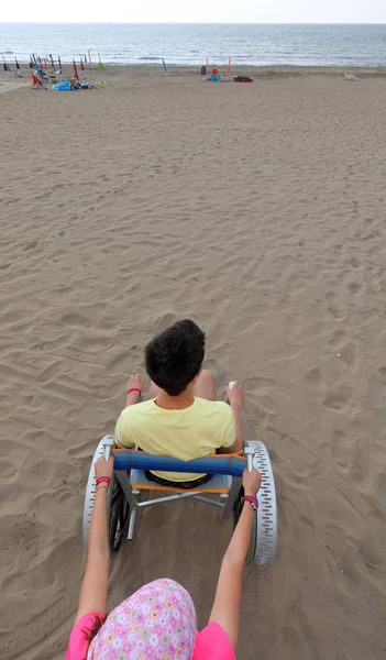 Bambina spinge una sedia a rotelle speciale sulla spiaggia di sabbia con — Foto Stock