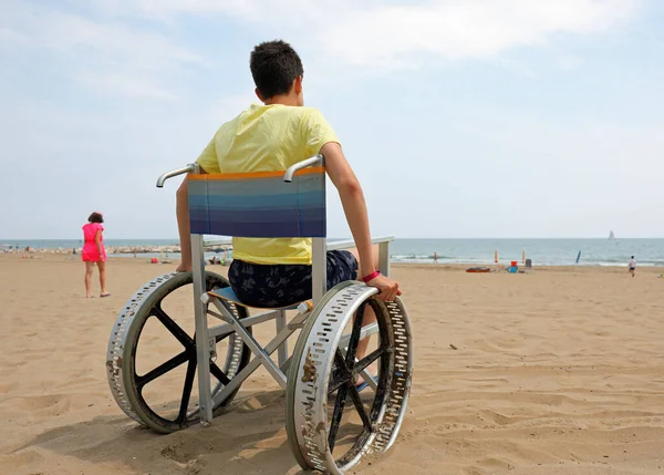 disabled boy on a wheelchair on the beach facing the sea