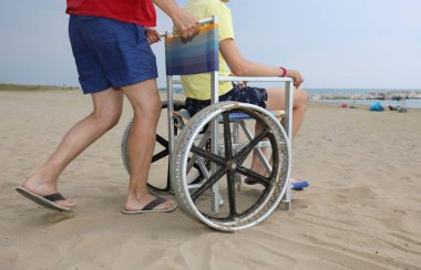 Caucasian Father pushes wheelchair with his son on sandy beach in summer