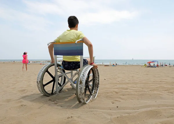 young boy in a wheelchair with large metal wheels to move easily on the sandy beach by the sea in summer