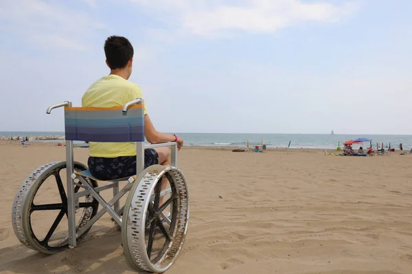 boy in a special wheelchair with very big metal wheels to move on the beach by the sea in summer