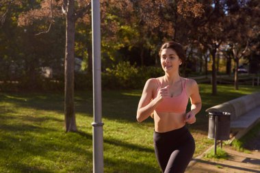 Kadın bir park bankının ve lamba direğinin yanından geçiyor.