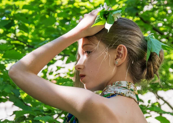 En ung flicka i en klänning med naken ryggar värt i lövverket i skogen håret inredda med bladverk. — Stockfoto