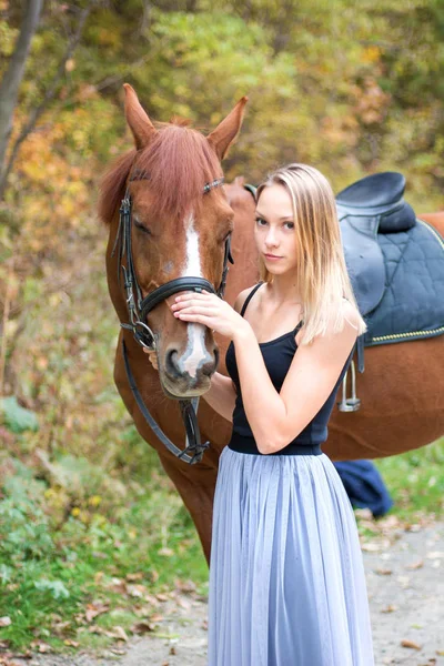 Egy fiatal, szőke lány pózol egy ló, egy gyönyörű lány, és egy erős ló. — Stock Fotó