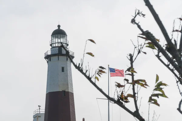 Lång vacker generiska fyren och amerikanska flaggan våg i havsbrisen vinden bakom kala hösten trädgren mot ljus vit himmel bakgrund — Stockfoto