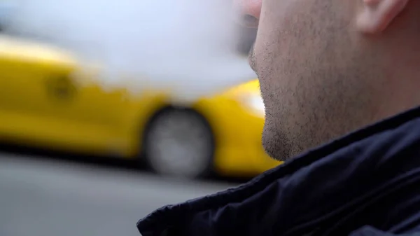 年轻的千禧年城市嬉皮士男子吹蒸汽电子香烟烟雾外的纽约市街头的白天时间。黄色出租车通过的背景。在肩部的视图 — 图库照片