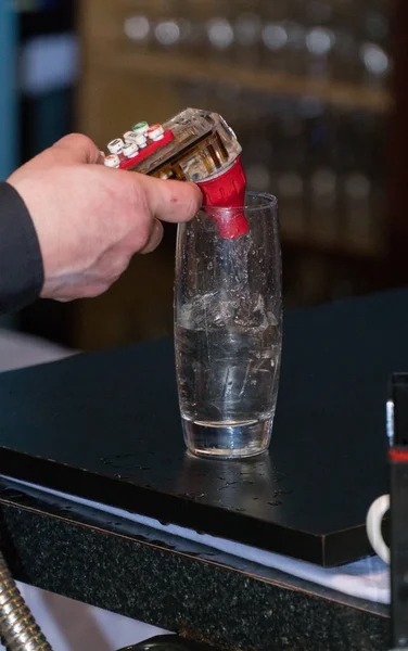 Bartender uses fountain soda gun to pour alcoholic mixed drink into tall glass with ice for thirsty customer.