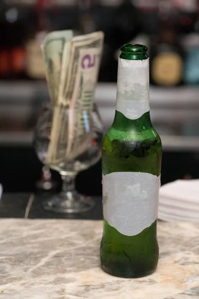 Generic beer bottle with blank label served by bartender waiting for customer to drink alcohol. Dollar bill money sits in brandy glass in background containing tips for restaurant server