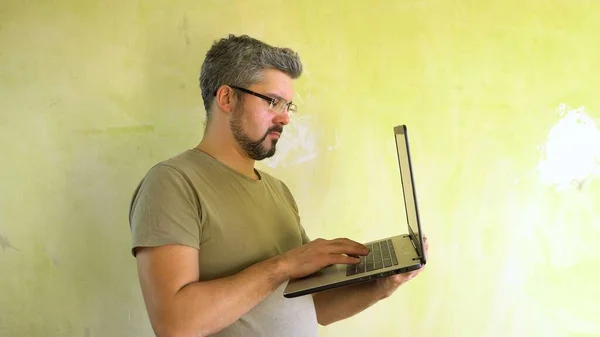 A man with glasses and a beard on his face. Holds a laptop in his hand. Types on the keyboard. Against the background of a green wall. Half-length portrait. Builder at the facility. Freelancer at home. Telework. Internet connection.