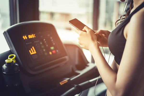 Азиатка, использующая смартфон при тренировке или силовых тренировках в фитнес-зале на беговой дорожке. Концепция расслабления и технологии. Спортивные упражнения и тема здравоохранения. Счастливое и комфортное настроение . — стоковое фото